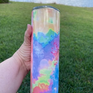 Puede incluir: Un vaso de acero inoxidable con un patr&oacute;n de tie-dye en tonos de rosa, azul, verde y amarillo. El vaso tiene una tapa plateada.