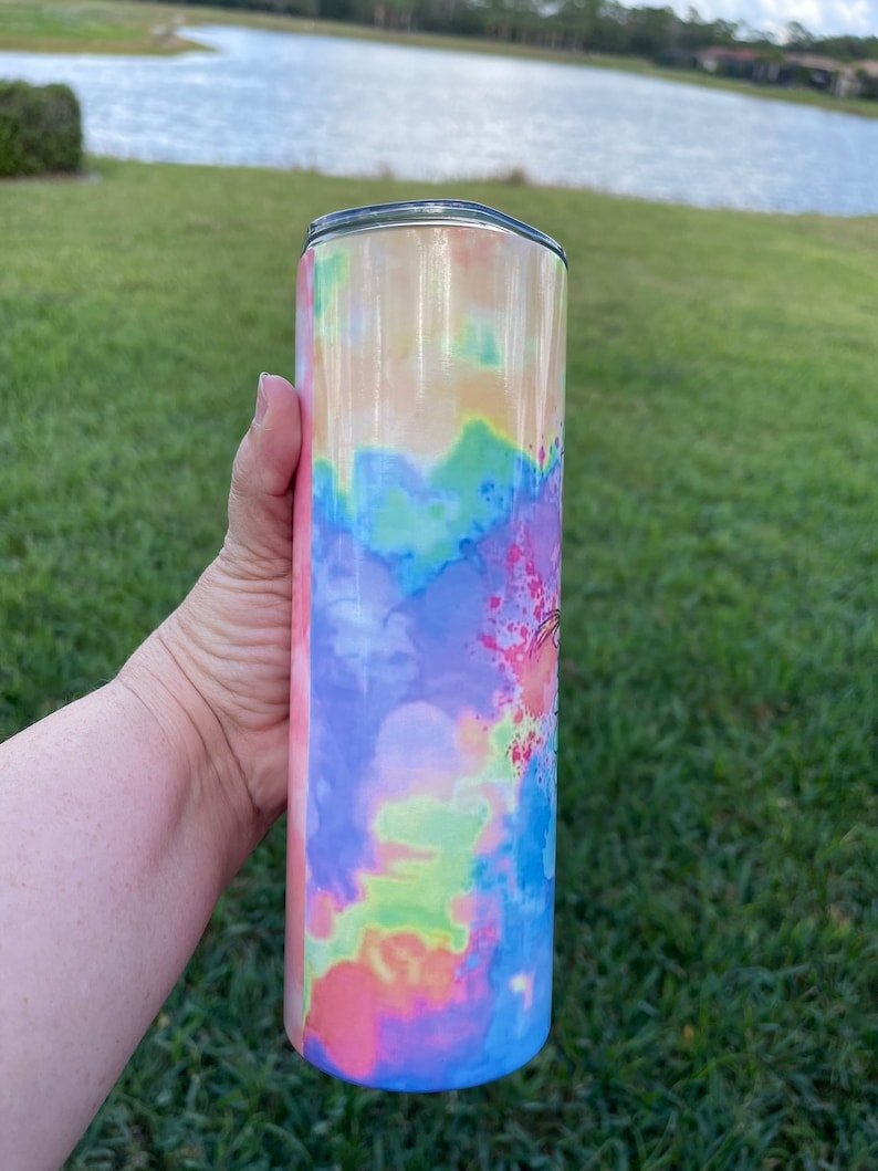 Puede incluir: Un vaso de acero inoxidable con un patr&oacute;n de tie-dye en tonos de rosa, azul, verde y amarillo. El vaso tiene una tapa plateada.