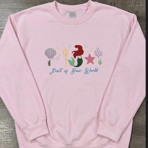 May include: A light pink crewneck sweatshirt featuring an embroidered design of a mermaid with red hair and a green tail, a gold trident, a purple seashell, a pink starfish, coral, and bubbles, with the text Part of Your World in blue script.