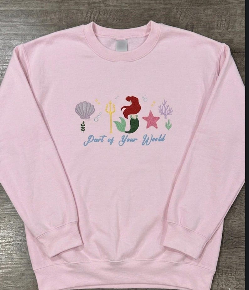 May include: A light pink crewneck sweatshirt featuring an embroidered design of a mermaid with red hair and a green tail, a gold trident, a purple seashell, a pink starfish, coral, and bubbles, with the text Part of Your World in blue script.