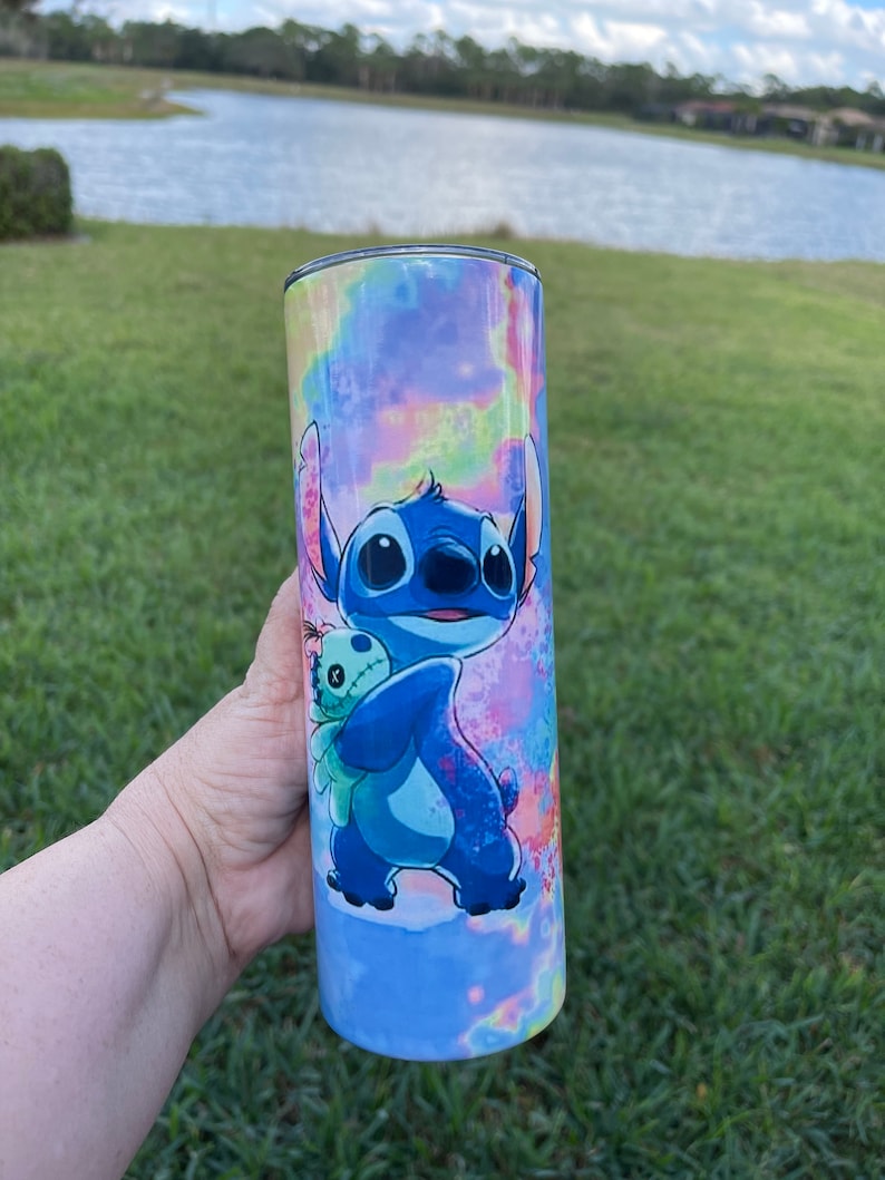 Puede incluir: Un vaso de acero inoxidable azul y morado con un dise&ntilde;o tie-dye y una imagen de dibujos animados de Stitch de Lilo & Stitch sosteniendo un juguete de peluche.