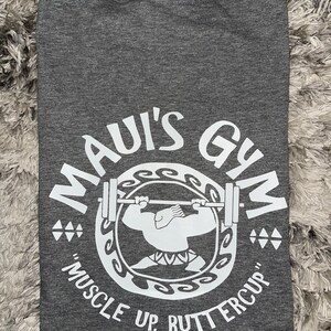 May include: Grey t-shirt with a white graphic of a cartoon character lifting weights. The graphic says "Maui's Gym" and "Muscle Up, Buttercup".
