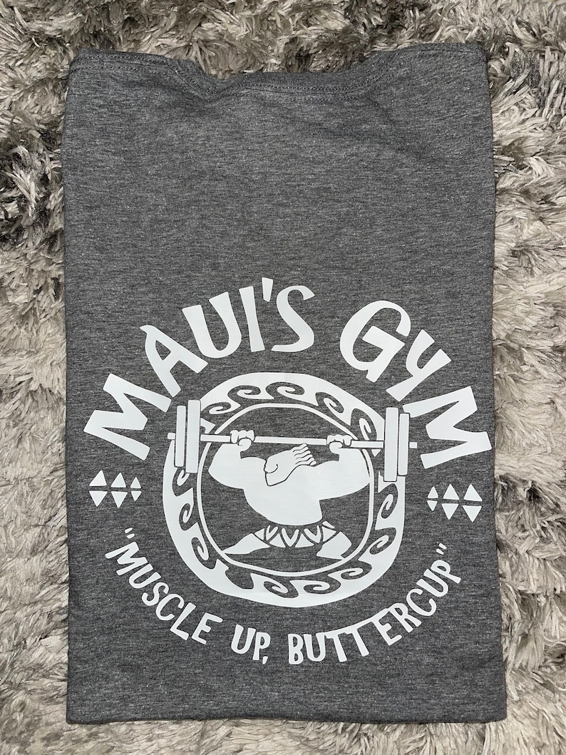 May include: Grey t-shirt with a white graphic of a cartoon character lifting weights. The graphic says "Maui's Gym" and "Muscle Up, Buttercup".