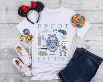 May include: White t-shirt with "EPCOT EST 1982" graphic, paired with blue jeans, white sneakers, and a sequined Minnie Mouse ear headband. Colorful lollipops add a playful touch to the scene.