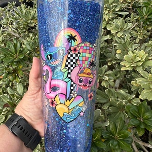 K&ouml;nnte beinhalten: Ein blauer Glitzerbecher mit einem durchsichtigen Deckel und einem Strohhalm. Der Becher zeigt ein farbenfrohes Design mit Stitch-Charakteren, einem Flamingo, einem Surfbrett, einem Regenbogen und einem Herz mit Wellen.