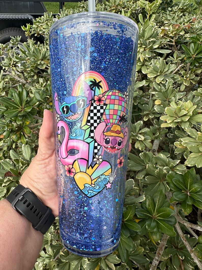 K&ouml;nnte beinhalten: Ein blauer Glitzerbecher mit einem durchsichtigen Deckel und einem Strohhalm. Der Becher zeigt ein farbenfrohes Design mit Stitch-Charakteren, einem Flamingo, einem Surfbrett, einem Regenbogen und einem Herz mit Wellen.