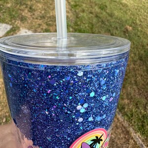 K&ouml;nnte beinhalten: Ein durchsichtiger Plastikbecher mit einer blauen Glitzerbasis und einem blauen und wei&szlig;en Cartoon-Charakter-Design. Der Becher hat einen durchsichtigen Deckel und einen Strohhalm.