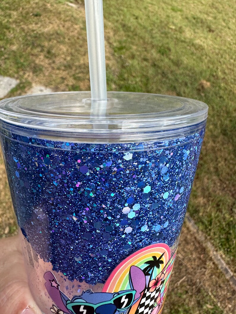 K&ouml;nnte beinhalten: Ein durchsichtiger Plastikbecher mit einer blauen Glitzerbasis und einem blauen und wei&szlig;en Cartoon-Charakter-Design. Der Becher hat einen durchsichtigen Deckel und einen Strohhalm.