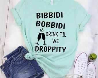 May include: A light green t-shirt with the text "Bibbidi Bobbidi Drink Til We Droppity" in black. The text is accompanied by a graphic of two champagne glasses with bubbles.