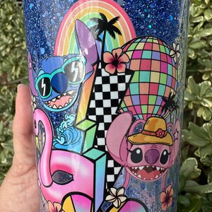 K&ouml;nnte beinhalten: Ein blauer Glitzerbecher mit einem bunten Design mit zwei Stitch-Charakteren, einem Regenbogen, Palmen, einem Hei&szlig;luftballon, einem Flamingo und einem karierten Muster.