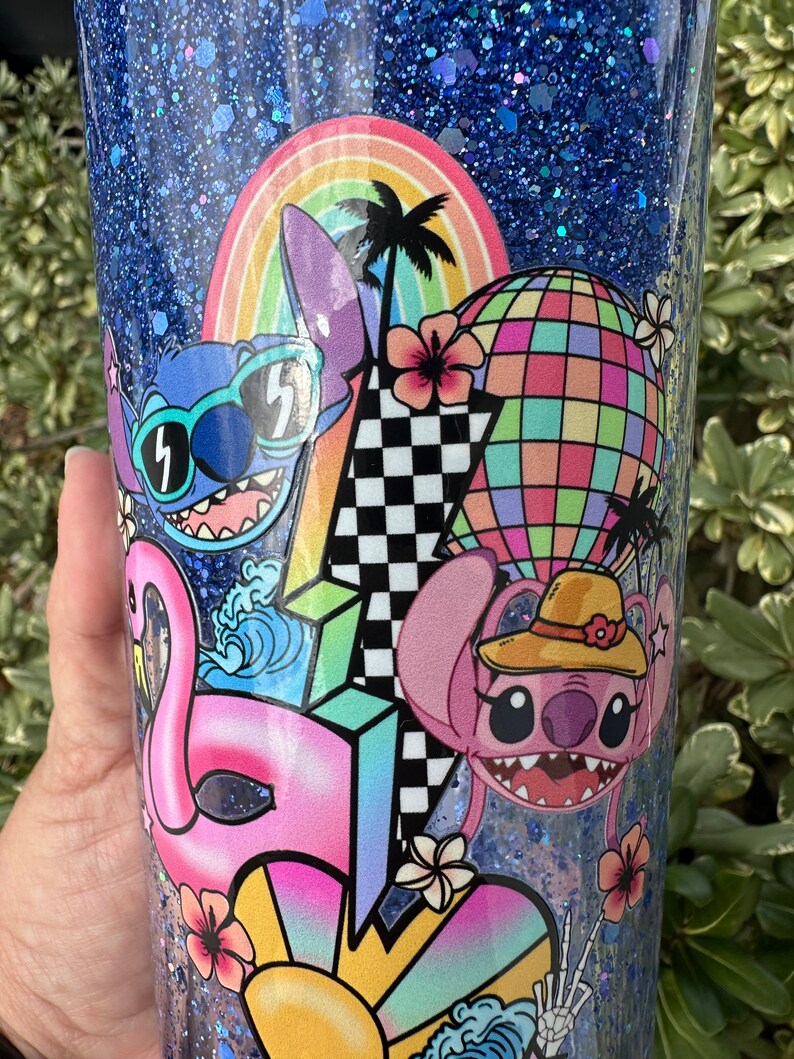K&ouml;nnte beinhalten: Ein blauer Glitzerbecher mit einem bunten Design mit zwei Stitch-Charakteren, einem Regenbogen, Palmen, einem Hei&szlig;luftballon, einem Flamingo und einem karierten Muster.
