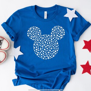 May include: A blue t-shirt with a white Mickey Mouse silhouette made of stars. The shirt is on a white surface with red, blue, and white stars scattered around it.