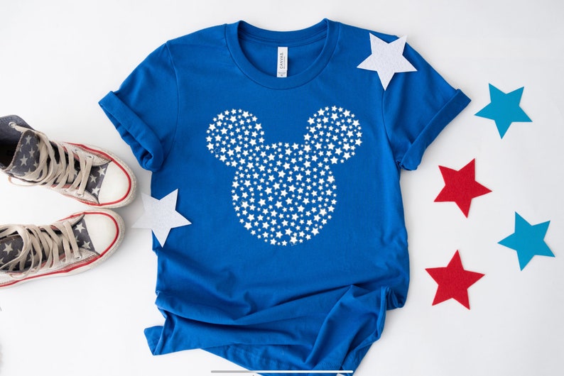 May include: A blue t-shirt with a white Mickey Mouse silhouette made of stars. The shirt is on a white surface with red, blue, and white stars scattered around it.