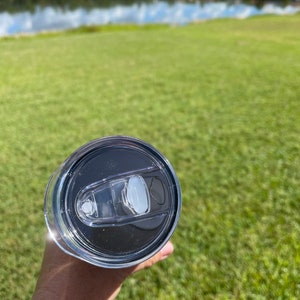 Puede incluir: Una taza de viaje de acero inoxidable con una tapa negra y un inserto de tapa de pl&aacute;stico transparente. La taza se sostiene en una mano contra un fondo borroso de hierba verde y un lago.