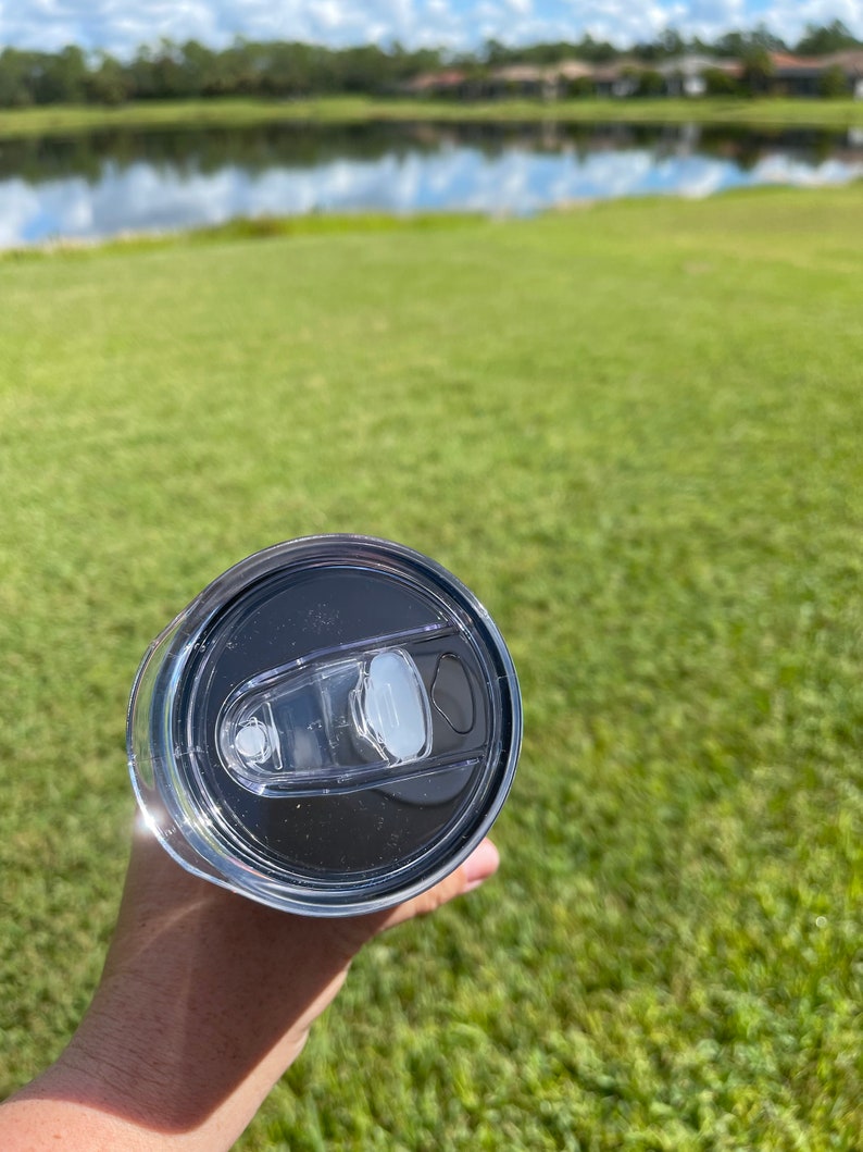 Puede incluir: Una taza de viaje de acero inoxidable con una tapa negra y un inserto de tapa de pl&aacute;stico transparente. La taza se sostiene en una mano contra un fondo borroso de hierba verde y un lago.