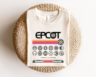 May include: A white t-shirt with a black graphic of the Epcot logo, a retro-style rainbow stripe, and various Epcot pavilion logos. The text "Community of Tomorrow" is printed below the logos. A black and white image of the Epcot monorail is printed at the bottom of the shirt.