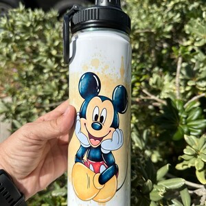 May include: White stainless steel water bottle with a black lid and a cartoon image of Mickey Mouse in a yellow and orange watercolor design.