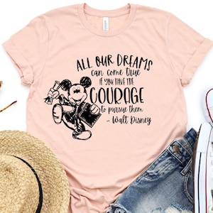 May include: A light pink t-shirt featuring a black and white illustration of Mickey Mouse holding a diploma and a book. The shirt has the quote "ALL OUR DREAMS can come true IF YOU HAVE THE COURAGE to pursue them - Walt Disney."