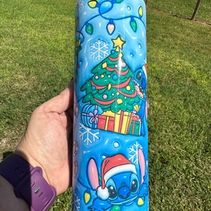 Może przedstawiać: Niebieski kubek termiczny ze stali nierdzewnej z świątecznym wzorem przedstawiającym Stitch'a z Lilo & Stitch. Wz&oacute;r zawiera choinkę, prezenty, płatki śniegu i lampki.