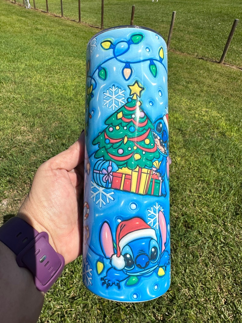 Może przedstawiać: Niebieski kubek termiczny ze stali nierdzewnej z świątecznym wzorem przedstawiającym Stitch'a z Lilo & Stitch. Wz&oacute;r zawiera choinkę, prezenty, płatki śniegu i lampki.