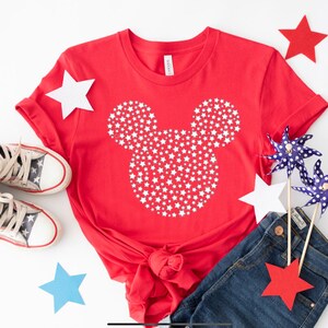 May include: Red t-shirt with a white Mickey Mouse silhouette made of stars. The shirt is paired with blue jeans, red and blue pinwheels, and red and blue stars.