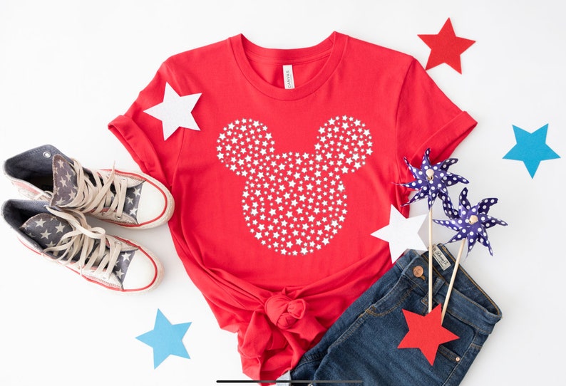 May include: Red t-shirt with a white Mickey Mouse silhouette made of stars. The shirt is paired with blue jeans, red and blue pinwheels, and red and blue stars.