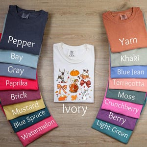 May include: A stack of t-shirts in various colors, including pepper, bay, gray, paprika, brick, mustard, blue spruce, watermelon, ivory, yam, khaki, blue jean, terracotta, moss, crunchberry, berry, and light green. The t-shirts are arranged in a vertical stack, with the ivory t-shirt in the center. The ivory t-shirt has a Thanksgiving-themed design with a pumpkin, a turkey, and other fall-themed elements.