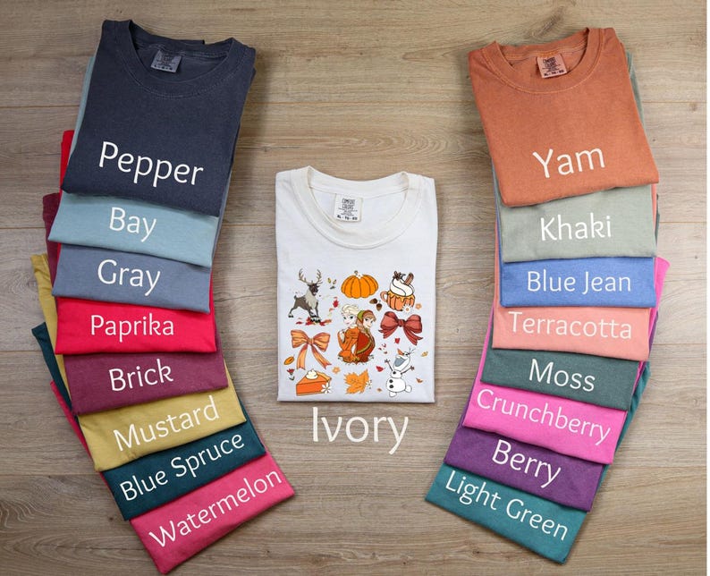 May include: A stack of t-shirts in various colors, including pepper, bay, gray, paprika, brick, mustard, blue spruce, watermelon, ivory, yam, khaki, blue jean, terracotta, moss, crunchberry, berry, and light green. The t-shirts are arranged in a vertical stack, with the ivory t-shirt in the center. The ivory t-shirt has a Thanksgiving-themed design with a pumpkin, a turkey, and other fall-themed elements.