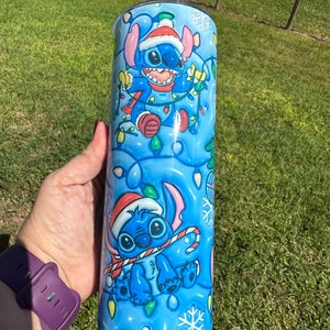 Może przedstawiać: Niebieski i biały kubek ze stali nierdzewnej z świątecznym wzorem przedstawiającym Stitch'a z Lilo & Stitch. Stitch ma na sobie czapkę Świętego Mikołaja i trzyma laskę cukrową. Tło jest niebieskie z płatkami śniegu i świątecznymi lampkami.