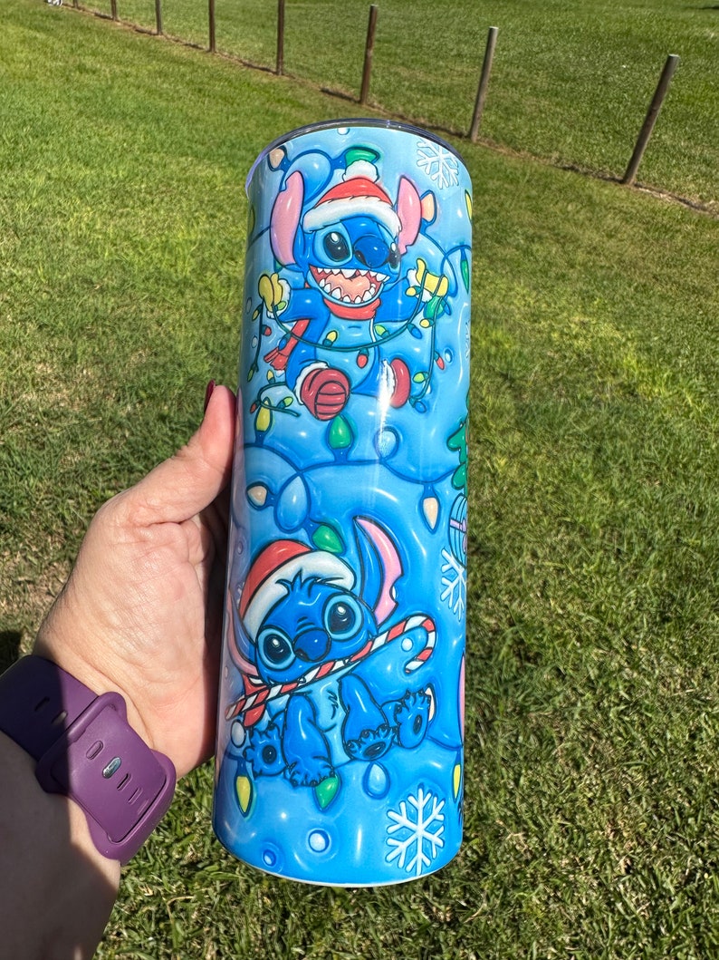 Może przedstawiać: Niebieski i biały kubek ze stali nierdzewnej z świątecznym wzorem przedstawiającym Stitch'a z Lilo & Stitch. Stitch ma na sobie czapkę Świętego Mikołaja i trzyma laskę cukrową. Tło jest niebieskie z płatkami śniegu i świątecznymi lampkami.