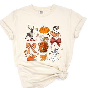 May include: A cream-colored t-shirt with a graphic print of Anna and Elsa from Frozen, a snowman, a reindeer, a pumpkin, a bow, a pie, and fall leaves.