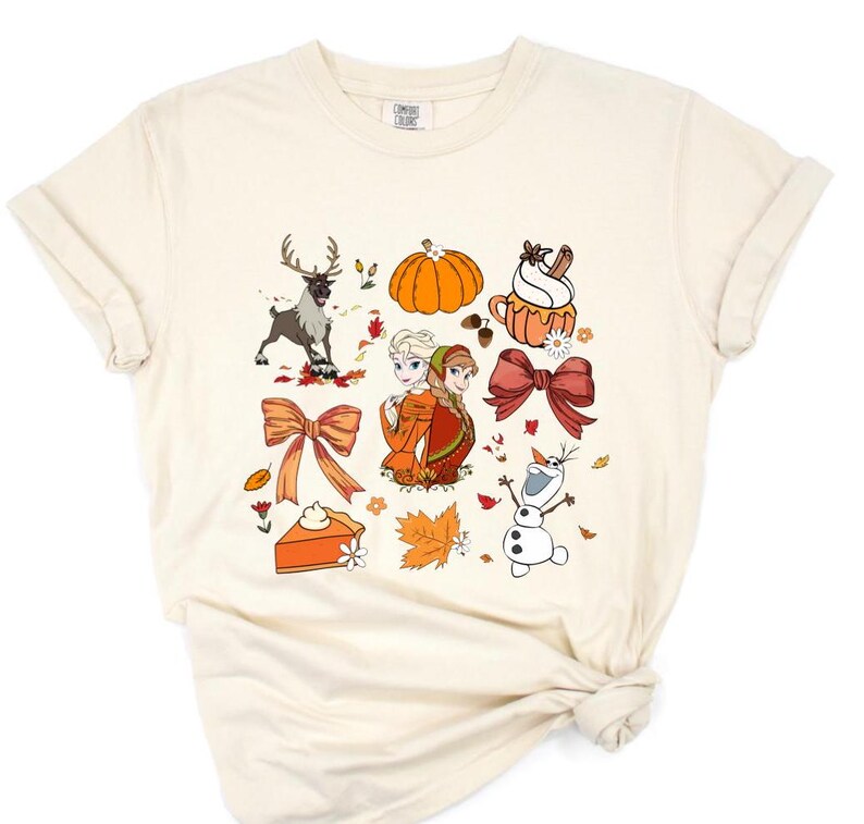 May include: A cream-colored t-shirt with a graphic print of Anna and Elsa from Frozen, a snowman, a reindeer, a pumpkin, a bow, a pie, and fall leaves.