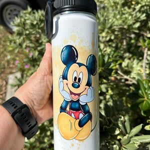 May include: White stainless steel water bottle with a black lid and a colorful image of Mickey Mouse. The image features Mickey Mouse sitting with his hands on his cheeks and a big smile. The background is a yellow and white watercolor design.