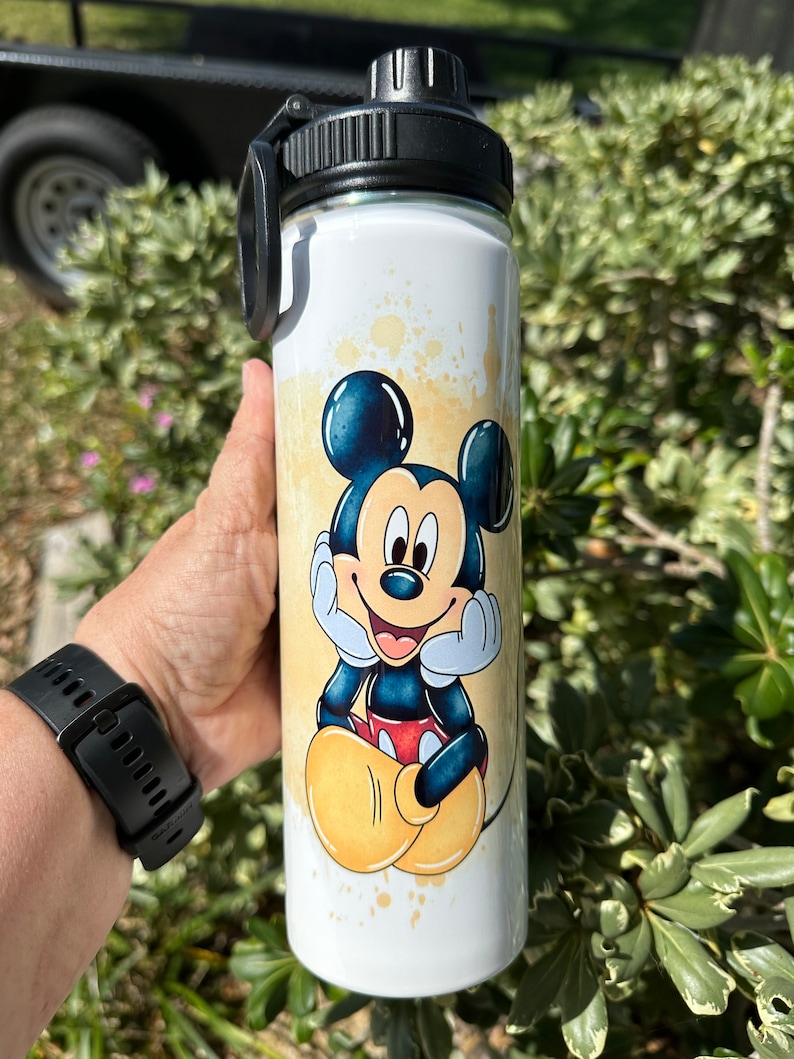 May include: White stainless steel water bottle with a black lid and a colorful image of Mickey Mouse. The image features Mickey Mouse sitting with his hands on his cheeks and a big smile. The background is a yellow and white watercolor design.