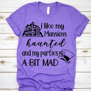 K&ouml;nnte beinhalten: Lilafarbenes T-Shirt mit schwarzem Text, der "Ich mag meine Herrenh&auml;user verflucht und meine Partys ein bisschen verr&uuml;ckt" lautet. Der Text wird von einer schwarzen Silhouette eines Spukhauses und einer schwarzen und wei&szlig;en Silhouette des Hutmachers begleitet.