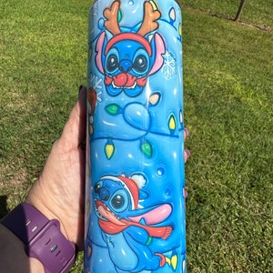 Może przedstawiać: Niebieski i biały kubek ze stali nierdzewnej z świątecznym wzorem przedstawiającym Stitch'a z Lilo & Stitch'a w czapce Świętego Mikołaja i rogach renifera. Kubek jest ozdobiony płatkami śniegu i świątecznymi lampkami.