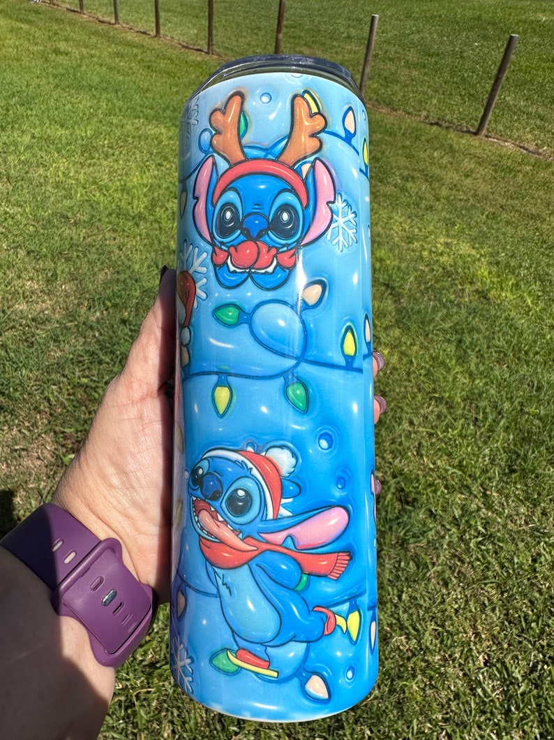 Może przedstawiać: Niebieski i biały kubek ze stali nierdzewnej z świątecznym wzorem przedstawiającym Stitch'a z Lilo & Stitch'a w czapce Świętego Mikołaja i rogach renifera. Kubek jest ozdobiony płatkami śniegu i świątecznymi lampkami.