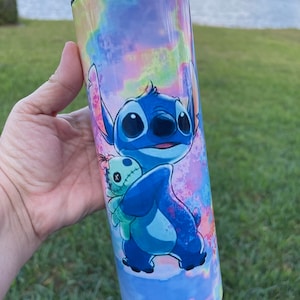 Puede incluir: Un vaso de acero inoxidable azul y verde con un fondo tie-dye que presenta a Stitch de Lilo & Stitch sosteniendo una peque&ntilde;a mu&ntilde;eca alien&iacute;gena verde.
