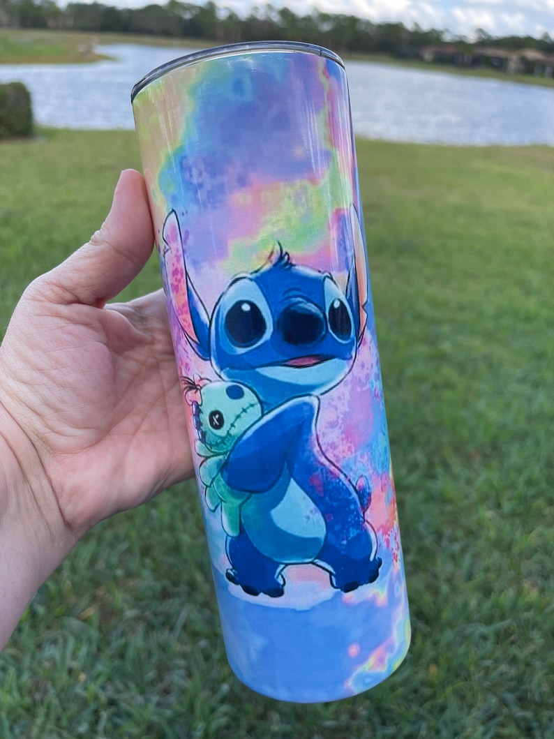 Puede incluir: Un vaso de acero inoxidable azul y verde con un fondo tie-dye que presenta a Stitch de Lilo & Stitch sosteniendo una peque&ntilde;a mu&ntilde;eca alien&iacute;gena verde.