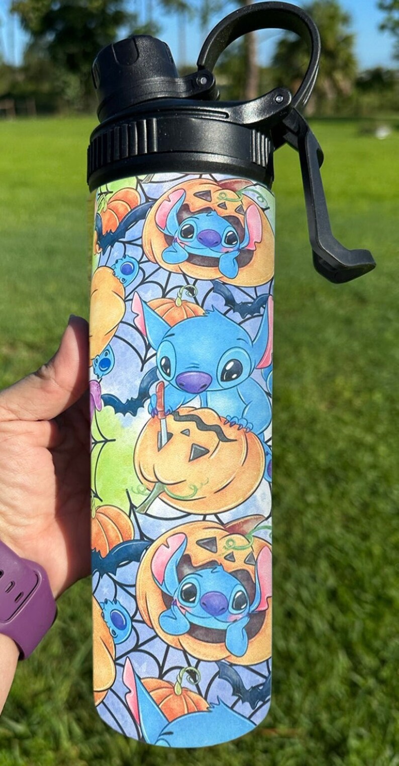 May include: A blue and white stainless steel water bottle with a black lid and a carrying handle. The bottle features a repeating pattern of Stitch from Lilo & Stitch carving pumpkins with bats and spiderwebs in the background.