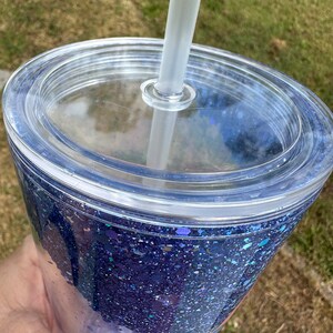 K&ouml;nnte beinhalten: Ein durchsichtiger Plastikbecher mit Deckel und Strohhalm. Der Becher ist mit blauem Glitzer gef&uuml;llt und hat ein Design auf dem Boden.