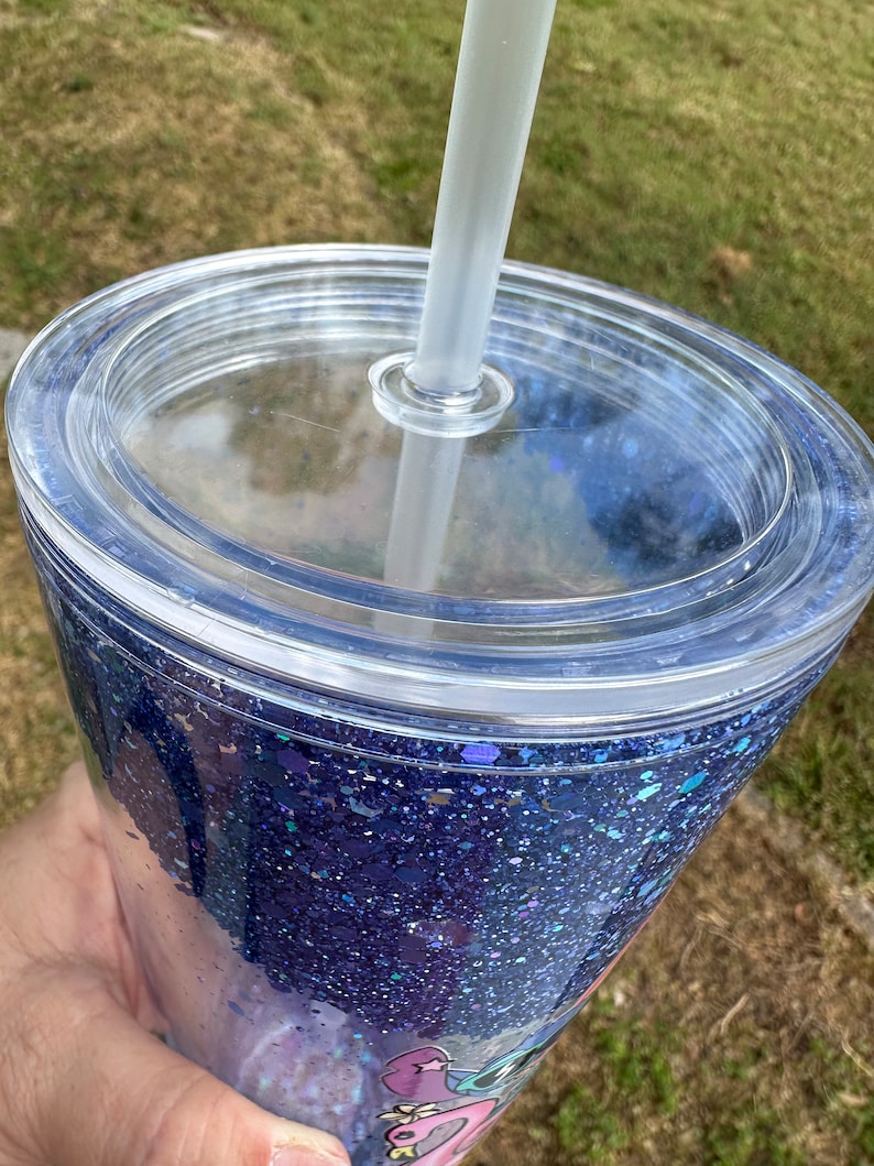 K&ouml;nnte beinhalten: Ein durchsichtiger Plastikbecher mit Deckel und Strohhalm. Der Becher ist mit blauem Glitzer gef&uuml;llt und hat ein Design auf dem Boden.