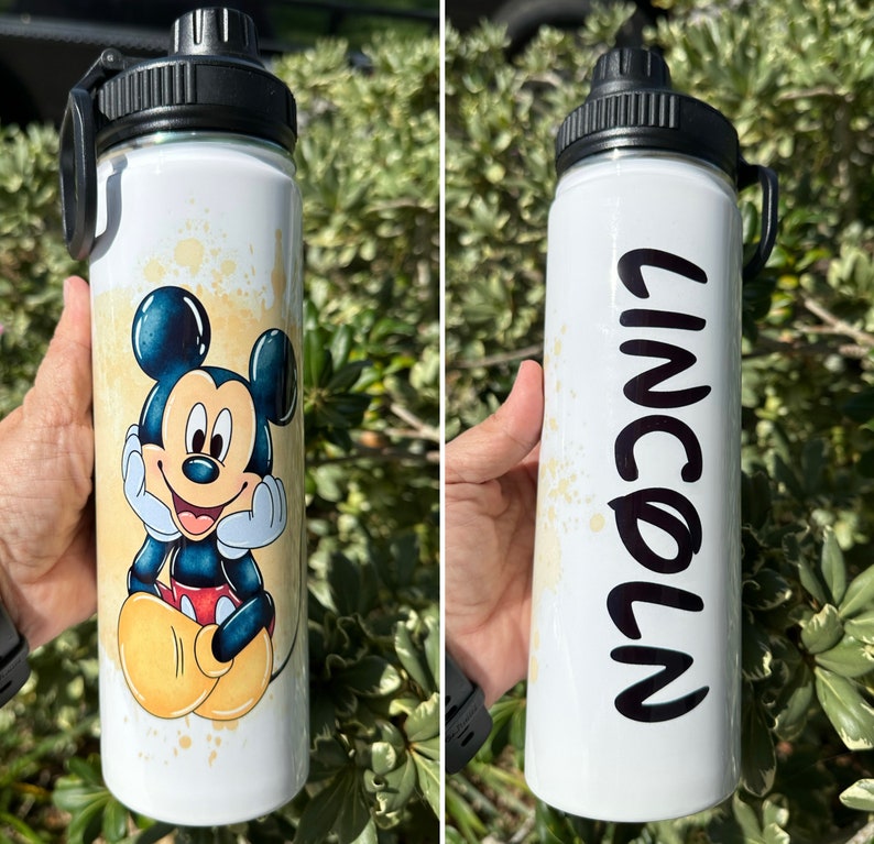 May include: Two white stainless steel water bottles with black lids and handles. One bottle features a cartoon image of Mickey Mouse. The other bottle has the name "Lincoln" written on it in black.