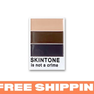May include: A white enamel pin with three horizontal stripes of colour: light brown, brown, and dark brown. The pin says "SKINTONE is not a crime".