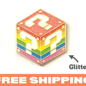 May include: A glittery, rainbow-colored question mark cube pin with gold trim. The pin features a white question mark on each side, set against a backdrop of rainbow stripes. The word "Glitter" is next to the pin.