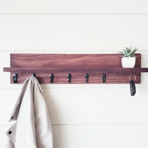 May include: A wooden wall mounted key rack with a shelf and six hooks. A beige jacket hangs on one of the hooks. A small potted plant sits on the shelf.