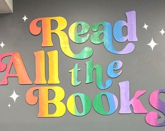 Puede incluir: Un letrero de pared con las palabras "Read All the Books" en colores arcoíris. Las letras están recortadas y superpuestas en una pared gris.