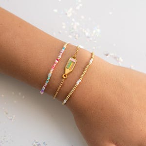 May include: Three delicate bracelets on a wrist. One features colorful beads, another a gold pencil charm, and the third has small gold beads. The bracelets are on a light background with scattered sparkles.
