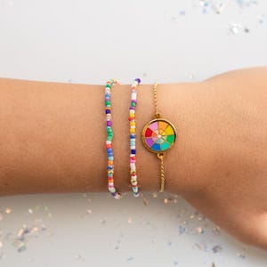 May include: Three bracelets on a wrist. Two are beaded, with colorful beads in various sizes. The third is a gold chain bracelet with a round pendant featuring a rainbow color wheel design. The bracelets are on a light skin tone.