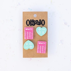 May include: Four pink and blue enamel pins with a calculator and heart design. The calculator pins say "I ♥ MATH" and the heart pins have math symbols. The pins are on a brown card with the word "아이방인" written on it.